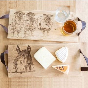 Zwei rechteckige Holzbretter mit braunen Lederschlaufen an den Seiten, liegend auf hellem Holztisch. Das obere Brett zeigt eine Gravur von drei Schafen und ist mit einem Glas Wasser und einem Glas Apfelsaft belegt. Das untere Brett zeigt eine Gravur eines