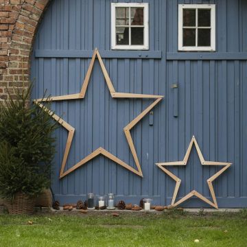 Holzsterne in zwei Größen lehnen an einer blauen Holzwand mit Fenstern. Daneben steht ein kleiner Tannenbaum im Topf. Auf dem Boden sind Kerzen in Gläsern und Tannenzapfen arrangiert.