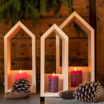 Drei rote Kerzen in holzgerahmten, hausförmigen Kerzenhaltern, umgeben von Tannenzapfen und Tannenzweigen auf einem Holztisch.