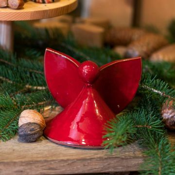 Rote Keramikfigur in Engelsform auf einem Holztisch, umgeben von Tannenzweigen und Nüssen. Der Engel hat stilisierte Flügel und einen runden Kopf ohne Gesicht.