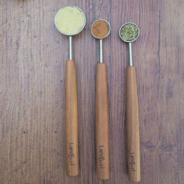 Drei Holzlöffel mit Gravur "Landlust", gefüllt mit unterschiedlichen Gewürzen, auf einer Holzoberfläche liegend. Unterschiedliche Löffelgrößen zeigen pulverisierte und getrocknete Kräuter. Schlichtes, natürliches Design.