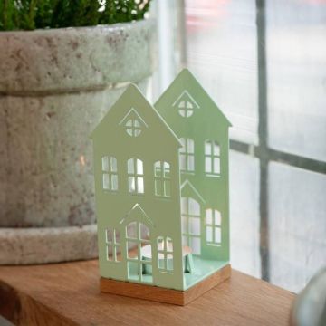 Pfefferminzgrüne Metall-Windlichtlaterne in Form eines Hauses mit mehreren Fensterausschnitten, auf einem Holzsockel; steht auf einem Holztisch neben einem großen Blumenübertopf.