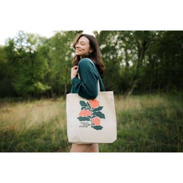 Eine Person im Freien trägt eine beigefarbene Stofftasche mit floraler Grafik und dem Text "Open eyes, open mind, open heart". Sie lächelt und steht vor Bäumen und Gras.