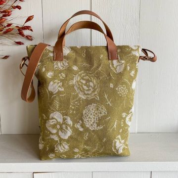 Olivegrüne Tasche mit weißen Blumenmustern und braunen Ledergriffen, daneben passende kleine Tasche mit Reißverschluss und Quasten. Taschen stehen vor hellholzwand auf Regal.