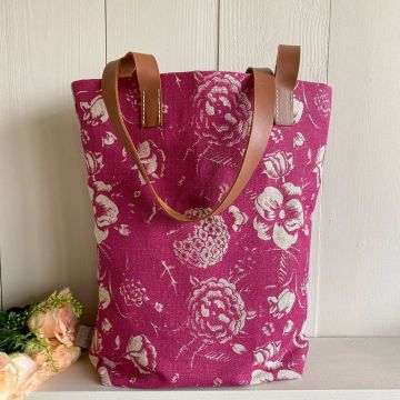 Tasche aus pinkem Stoff mit cremefarbenem Blumenmuster, zwei braune Lederhenkel, steht auf hellem Untergrund vor weißer Holzrückwand, links daneben liegen rosafarbene Kunstblumen.