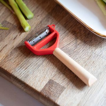 Gemüseschäler mit rot-schwarzem Kunststoffkopf und Holzgriff liegt auf einem Holzbrett; im Hintergrund angeschnittene grüne Spargelstangen und ein weißer Teller sichtbar.
