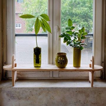 Holzregal auf Fensterbank mit drei Pflanzen in verschiedenen Töpfen, darunter ein Glasgefäß. Hintergrund zeigt Fensterblick ins Freie.