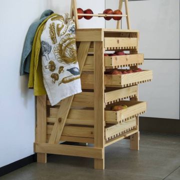 Holzregal mit vier offenen Schubladen, gefüllt mit roten Äpfeln. Auf der linken Seite hängt ein bunt bedrucktes Geschirrtuch. Das Regal steht auf einem Fliesenboden neben einer weißen Wand.