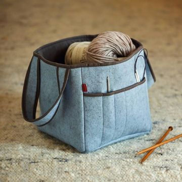 Hellblaue Filztasche mit braunem Rand, steht auf hellem Teppich; enthält ein Wollknäuel, eine Banderole mit Aufschrift „Wool“ sowie Strick- und Häkelnadeln in Außentaschen; zwei Holznadeln liegen daneben.