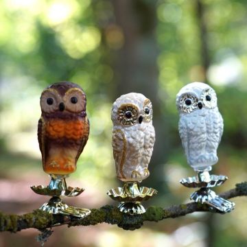 Drei dekorative Eulenfiguren in verschiedenen Farben stehen auf einer Astkerze, jede montiert auf einem metallenen Clip. Der Hintergrund ist ein verschwommener Wald.