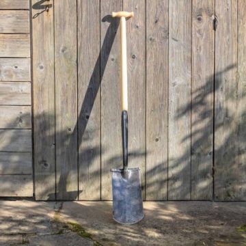 Garten-Spaten mit T-Griff aus hellem Holz und schwarzem Metallblatt, aufrecht an eine Holzwand gelehnt, vollständig sichtbar, auf Steinboden stehend, fotografiert bei Tageslicht.