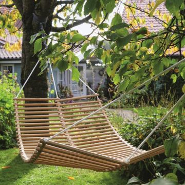 Holzlatten-Hängematte, an Baumästen befestigt, über grünem Rasen im Garten. Im Hintergrund steht ein Haus mit blauen Wänden und Ziegeldach, umgeben von Pflanzen und Sträuchern. Sonniges Wetter, ruhige Atmosphäre.