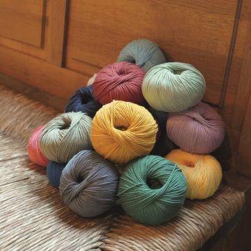 Mehrfarbige Wollknäuel in verschiedenen Farben, darunter Gelb, Blau, Rot und Grün, liegen gestapelt auf einem geflochtenen Sitz. Sie sind rundlich geformt und zeigen eine weiche Textur, geeignet zum Stricken oder Häkeln.