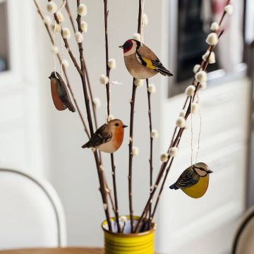 Zweige in gelber Vase mit weißen Knospen und vier hängenden Vogelholzdekorationen in verschiedenen Farben und Designs.