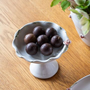 Weiße Porzellanschale auf Sockel mit sieben dunklen Pralinen, arrangiert auf einem Holztisch. Daneben sind Teile einer dekorierten Vase mit grünen und rosafarbenen Blumen sichtbar.