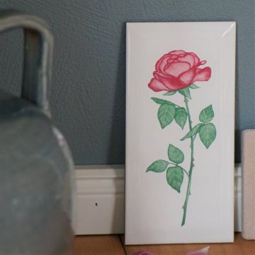 Weiße Keramikfliese mit handgemaltem roten Rosendekor, stehend vor einer grauen Wand auf einem Holzfußboden, neben einem angedeuteten grauen Objekt links und einem weiteren Objekt rechts.
