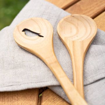 Zwei hölzerne Kochlöffel liegen auf einem grauen Leinenhandtuch auf einem Holztisch. Einer der Löffel hat eine ausgeschnittene Form in Silhouette eines Vogels.