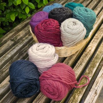 Verschiedene Wollknäuel in einem Korb auf einer Holzbank. Die Farben umfassen Rot, Blau, Lila, Grün, Schwarz, Beige und Weiß. Die Wollknäuel sind dicht nebeneinander angeordnet und teilweise außerhalb des Korbes platziert.