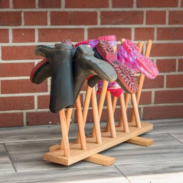 Holz-Schuhregal mit mehreren Stangen, auf denen verschiedene Arten von Schuhen, darunter schwarze Gummistiefel und rosa Clogs, stehen. Hintergrund aus roten Backsteinen.