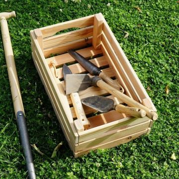 Holzkiste mit Gartengeräten (Hacke, Schaufel, Spaten) auf Rasen. Links ist ein weiteres langes Gartengerät. Die Kiste hat Seitengriffe und ist aus hellem Holz, die Werkzeuge haben Holzgriffe und metallene Köpfe.