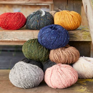 Mehrere verschiedenfarbige Wollknäuel, darunter Rot, Dunkelblau, Goldbraun, Grün, Rosa, Beige, Grau und Schwarz, liegen dekorativ gestapelt auf alten Holzstufen; das Bild dient der Produktdarstellung in einem Online-Shop.