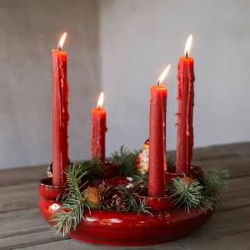 Ein roter Adventskranz mit vier brennenden roten Stabkerzen, geschmückt mit Tannenzweigen und kleinen Zapfen, steht auf einem Holztisch. Wachs ist an den Kerzen heruntergelaufen, was eine warme, festliche Atmosphäre schafft.