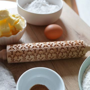 Holz-Nudelholz mit eingraviertem Hasenmotiv, umgeben von Zutaten wie Butter, Mehl, Ei und Zimt, auf einem Holztisch.