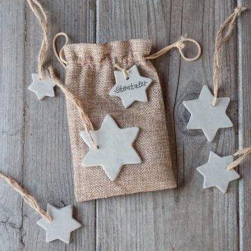Ein Jutebeutel mit dem Aufdruck "Sterntaler" liegt auf einem Holztisch. Um den Beutel herum liegen mehrere sternförmige Anhänger aus Keramik mit Schnüren.