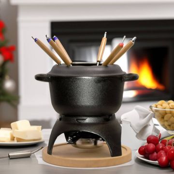 Fondue-Set aus schwarzem Metall mit Holzgabeln, platziert auf einem Tisch vor einem Kamin. Im Hintergrund weihnachtliche Dekoration. Auf dem Tisch ein Teller mit Butter und ein weiterer mit Trauben und Macadamianüssen.
