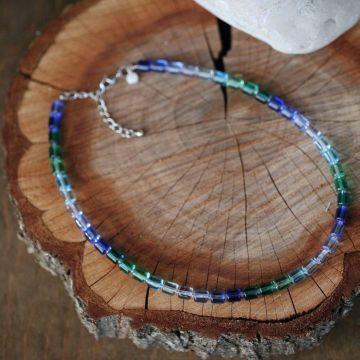 Halskette aus rechteckigen, durchsichtigen Glasperlen in Blau- und Grüntönen, auf Holzhintergrund liegend. Silberner Verschluss an einer Metallkette.