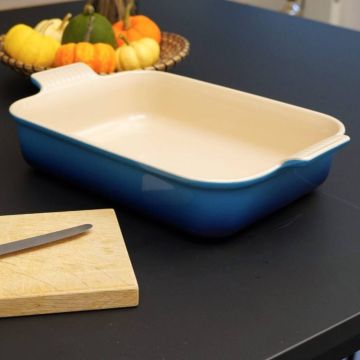 Leere, rechteckige Auflaufform aus Keramik mit blauer Außenseite auf einem schwarzen Tisch. Im Hintergrund ein Korb mit Zierkürbissen, im Vordergrund ein Holzbrett mit einem Messer.