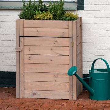 Hölzerner Pflanzkübel mit Kräutern, quadratisch, vor weißer Ziegelwand stehend. Rechts daneben eine grüne Gießkanne auf rotem Backsteinboden.