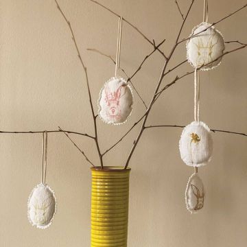 Gelber Keramikvase mit kahlen Zweigen, daran befestigt ovale Stoffanhänger mit gestickten Tiermotiven in verschiedenen Farben.