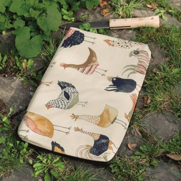 Laptop-Hülle mit buntem Hühner-Muster auf Gras und Pflastersteinen liegend; im Hintergrund ein Holzgriff-Werkzeug.