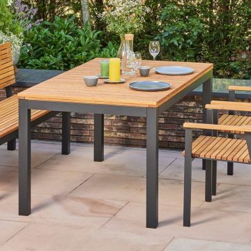 Gartenmöbel-Set auf Terrasse mit Holztisch und Stühlen auf Metallrahmen, dekoriert mit Gläsern, Tellern, bunten Kerzen und einer Glasflasche. Im Hintergrund grüne Sträucher und Pflanzen.