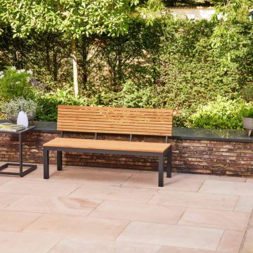 Holzbank mit Metallfüßen auf einer gepflasterten Terrasse, umgeben von Backsteinmauer und grünen Büschen im Hintergrund. Links daneben steht ein kleiner Tisch mit Deko-Objekten.