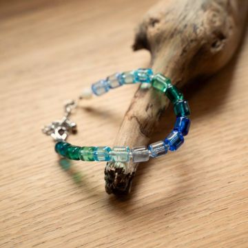Armband aus grünen, blauen und klaren quadratischen Perlen auf hellem Holztisch, drapiert über ein Stück Treibholz.