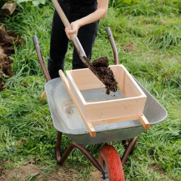 Person schaufelt Erde durch ein Holzsieb in eine Schubkarre auf einer Wiese.