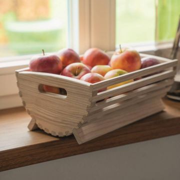 Holzkiste mit Griffen auf beiden Seiten, gefüllt mit roten Äpfeln, steht auf einem Fenstersims vor einem Fenster mit unscharfer Außensicht.