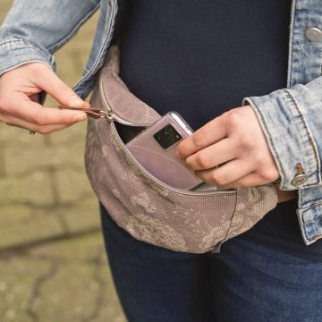 Person hält ein Smartphone und öffnet eine Hüfttasche mit floralem Muster. Die Person trägt eine Jeansjacke. Das Smartphone ist teilweise sichtbar, während es in die Tasche gesteckt wird.