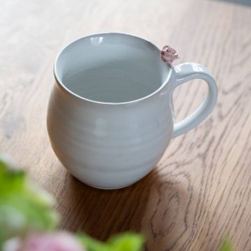 Weiße keramische Tasse mit runden Form, auf Tisch aus Holz. Kleines rosafarbenes Dekoelement an Tassenrand. Im Vordergrund unscharfe grüne und rosa Pflanzen.