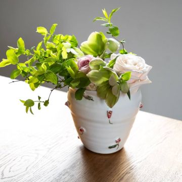 Weiße Vase mit roten Rosenmustern, gefüllt mit grünen Blättern und hellen Rosen auf einem Holztisch, vor neutralem Hintergrund.