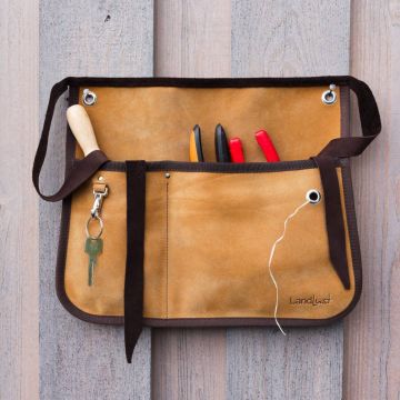 Eine braune Lederwerkzeugtasche mit mehreren Fächern, hängt an einer Holzplatte. Enthält Werkzeuge wie Schraubenzieher und Zangen. Ein Schlüsselbund mit Schlüssel ist an der linken Seite befestigt. Aufdruck: "Landlust".