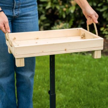 Holztablett mit zwei Seilgriffen wird von Händen gehalten; grüne Grasfläche und Pflanzen im Hintergrund, Personenbeine in Jeans sichtbar.