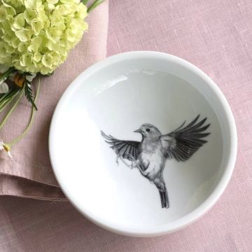 Weiße Schale mit Schwarz-Weiß-Abbildung eines fliegenden Vogels im Inneren, auf rosa Stofftuch platziert. Links oben befinden sich gelbe Blüten und grüne Blätter.