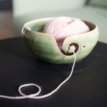 Grüne Keramikschale mit geschwungener Öffnung, hält rosa Wollknäuel. Wolle zieht durch seitlichen Schlitz, liegt auf schwarzer Oberfläche. Ideal zum ordentlichen Abwickeln beim Stricken oder Häkeln.