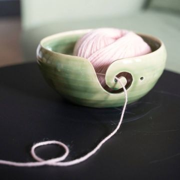 Grüne Keramikschale mit geschwungener Öffnung, hält rosa Wollknäuel. Wolle zieht durch seitlichen Schlitz, liegt auf schwarzer Oberfläche. Ideal zum ordentlichen Abwickeln beim Stricken oder Häkeln.