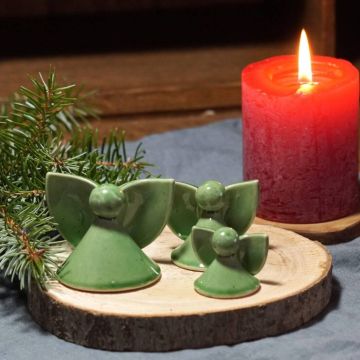Drei grüne Keramikfiguren in Engelsform auf einer runden Holzscheibe, neben einem Zweig und einer brennenden roten Kerze, in gemütlicher Umgebung arrangiert.
