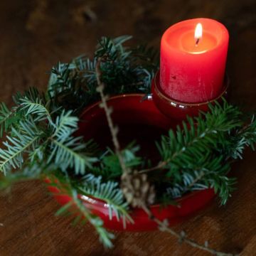 Eine brennende rote Kerze steht in einem roten Kerzenhalter, dekoriert mit Tannenzweigen und einem kleinen Zapfen. Der Kerzenhalter befindet sich auf einem Holztisch und verleiht der Szene eine warme, festliche Atmosphäre.