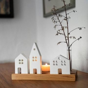 Drei weiße Keramikhäuschen mit Fensterausschnitten stehen auf einem Holzsockel, dahinter brennt ein Teelicht; rechts daneben steht ein Glas mit einem Zweig. Die Szene befindet sich auf einer Holzoberfläche vor heller Wand.
